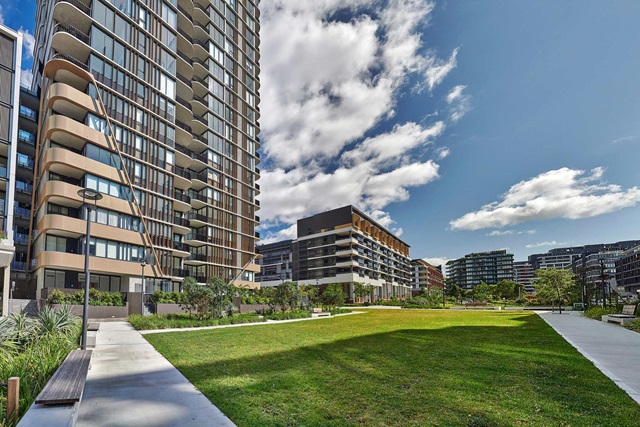 Modern apartment buildings surround a landscaped green lawn with pathways, benches, and scattered trees under a partly cloudy sky.