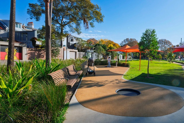 A person with a stroller walks along a path in a park with benches, greenery, and orange shade structures on a sunny day.