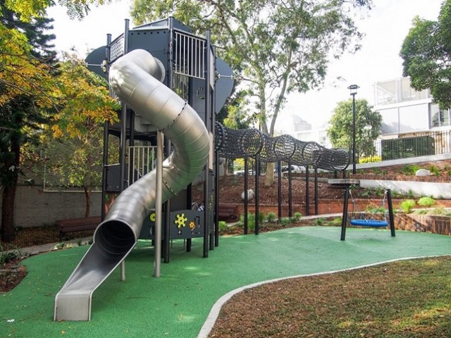 Playground with a tall metal slide, climbing structure, and swings set on a green rubber surface surrounded by trees and grass.