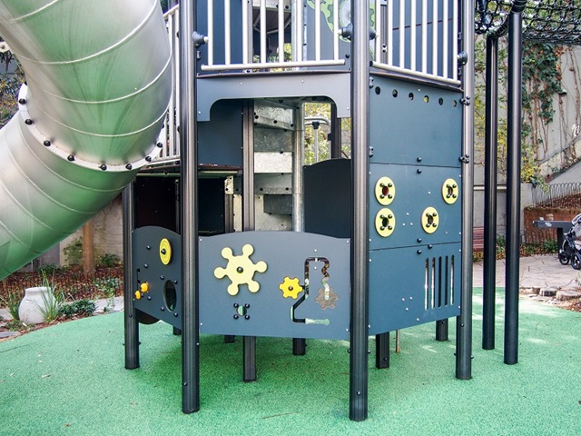 Playground structure with a metal slide, green flooring, and decorative panels featuring yellow gears and shapes.