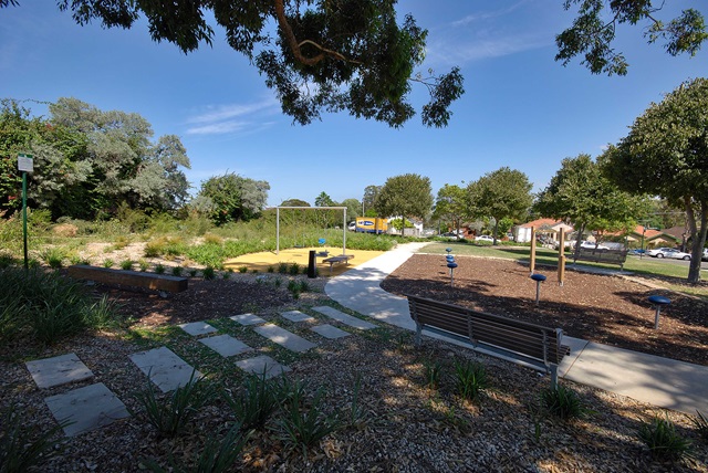 Park with paved paths, a bench, playground equipment, and trees. A swing set is visible, surrounded by mulch and grass. Blue sky and distant buildings in the background.