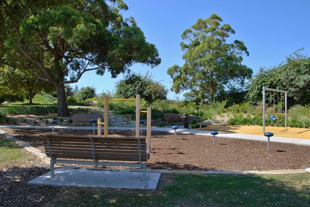 Outdoor fitness area with exercise equipment, wooden bench, and trees in a park setting under a clear blue sky.