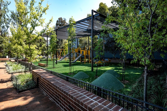 Playground with slides and climbing equipment on a grassy area, surrounded by trees and brick pathways, under a metal pergola.