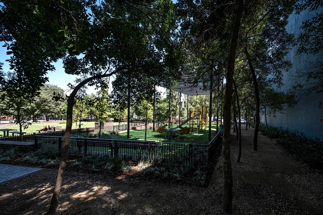 A fenced playground with a slide is surrounded by trees and grass in an open park area.