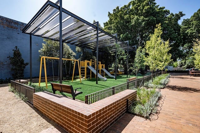 Playground with yellow swings and slides, shaded by a metal structure, surrounded by a brick border and greenery, next to a paved area.