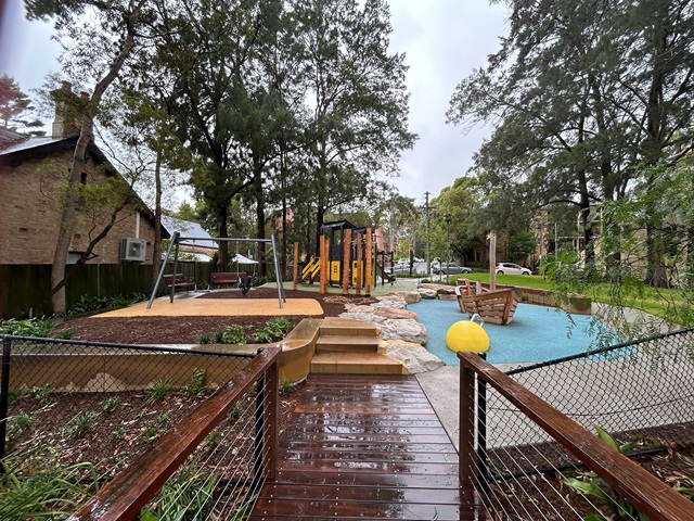 A playground with swings, a climbing structure, and a slide is surrounded by trees. A wooden bridge and sandy play area are visible. The sky is overcast.