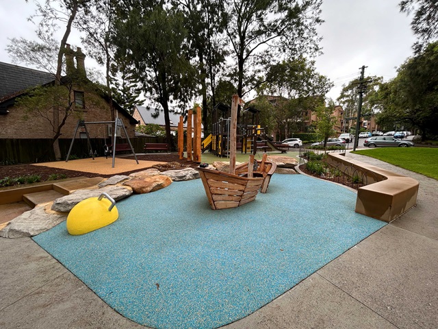 A playground with a wooden boat structure, swings, climbing equipment, and a yellow play object on a blue surface, surrounded by trees and benches.