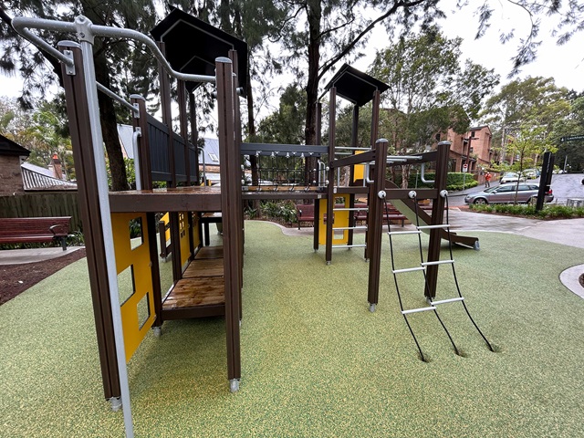 A children's play structure with climbing features, slides, and platforms on a textured ground. Trees and benches are in the background.