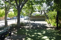 A playground with a single swing set under the shade of large trees. A fence borders the area, and a quiet residential street is visible in the background.