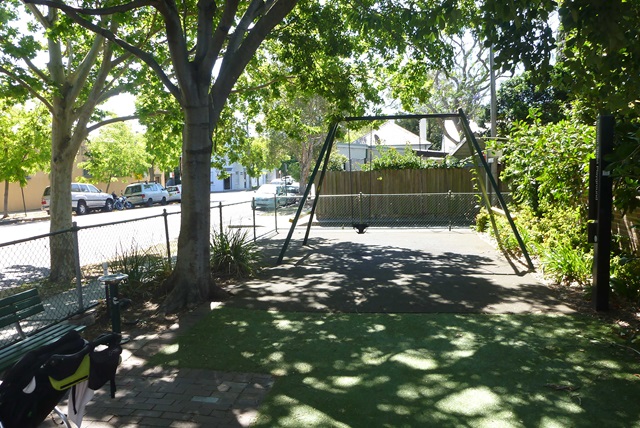 A playground with a single swing set under the shade of large trees. A fence borders the area, and a quiet residential street is visible in the background.