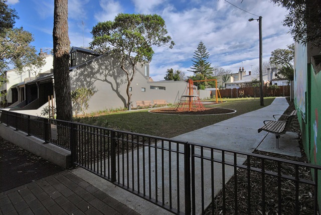 A fenced playground with a climbing structure and benches, set in a courtyard surrounded by trees and houses under a cloudy sky.