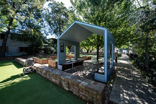 A small open pavilion with benches in a park setting, surrounded by trees and a brick walkway. A stroller is parked nearby on a patch of green artificial turf.