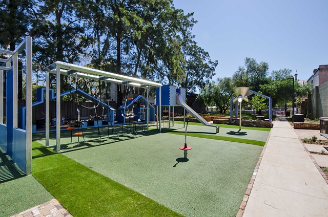 A playground with swings, slides, climbers, and artificial grass. Trees and benches are in the background. A pathway runs alongside the play area.