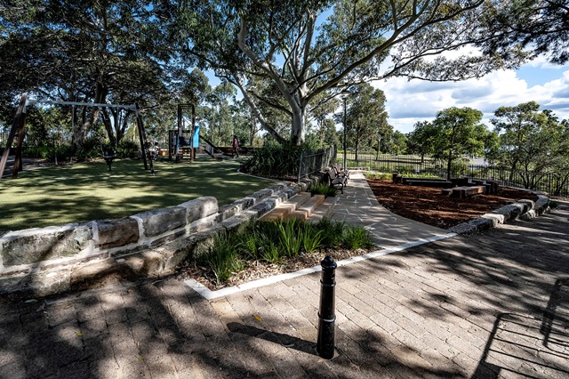 A park with a swing set, a slide, and a paved walkway surrounded by trees and greenery under a partly cloudy sky.