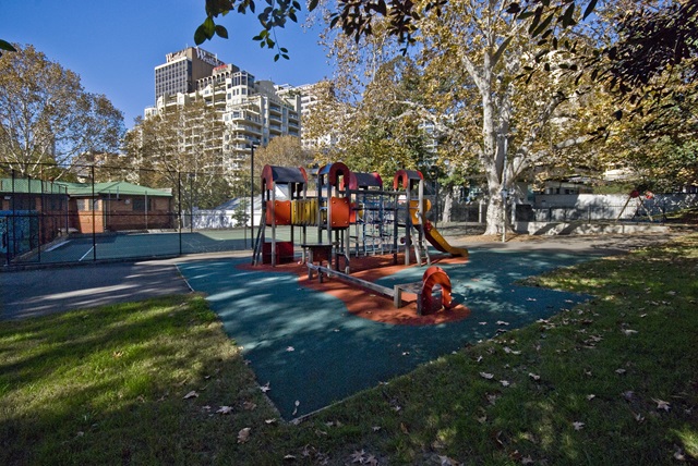 Playground with slides and climbing structures surrounded by trees, adjacent to a fenced court, with tall buildings in the background under a clear sky.