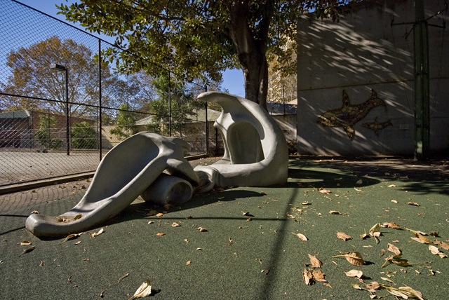 Two abstract concrete sculptures in a park setting with leafy ground and a fenced area in the background.