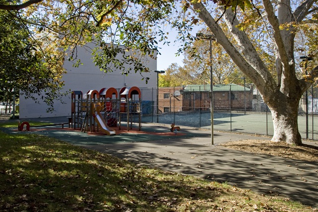 A playground with slides and climbing frames is adjacent to a fenced tennis court. Large trees provide shade, and fallen leaves cover the ground.