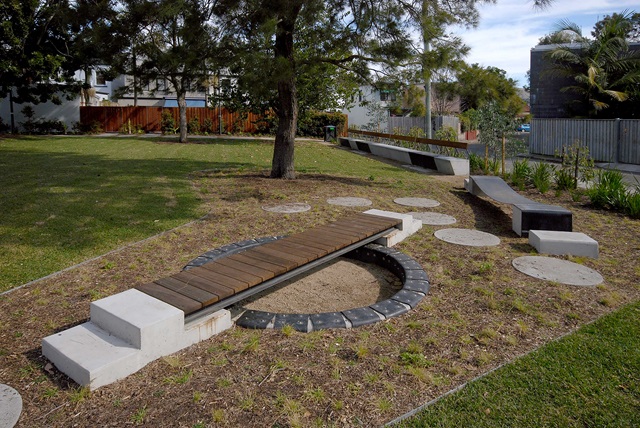 Wooden bridge over a circular sandpit with nearby stepping stones and modern benches in a grassy park setting.