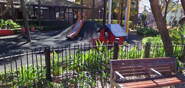 A playground with a slide on a mound, a small playhouse, and a bench in the foreground, surrounded by greenery and a fence.