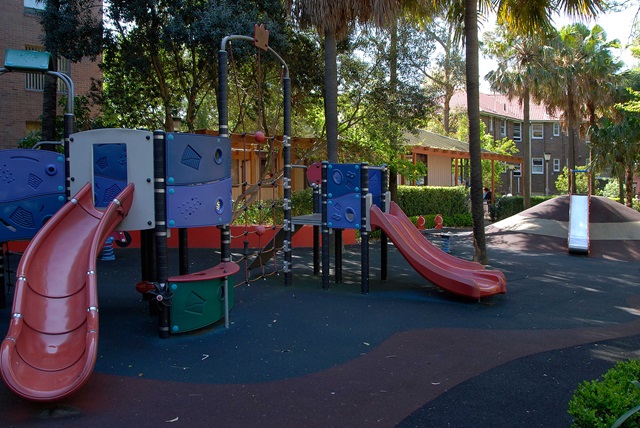 Playground with slides, climbing structures, and a rubber surface, surrounded by greenery and buildings.