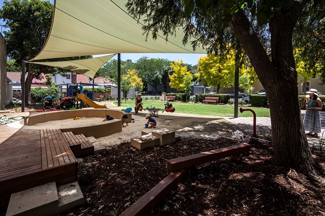 Children play in a shaded playground with slides and sandboxes, surrounded by trees and benches. Adults stand nearby, observing.