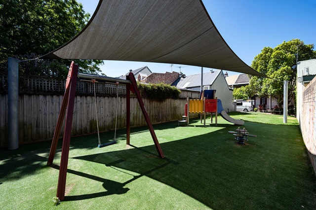 Playground with a swing set, slide, and climbing structure under a large shade sail on artificial grass, surrounded by a wooden fence and buildings.