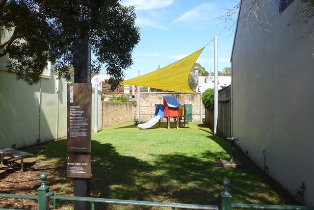 Small urban playground with a slide and climbing structure under a yellow sunshade. Surrounded by walls and a tall tree on the left. Informational sign at the entrance.