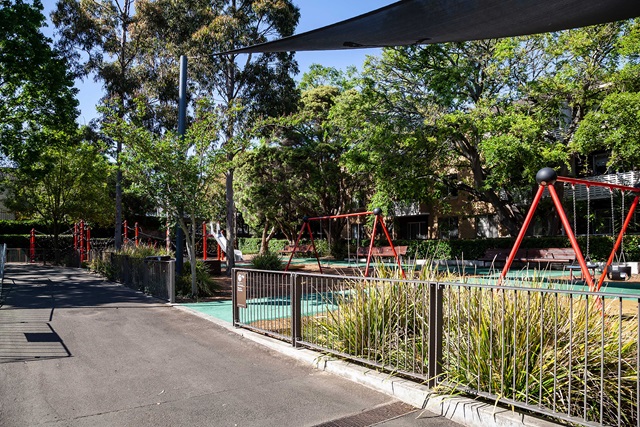Playground with swings and climbing equipment, surrounded by trees and greenery, adjacent to a paved pathway on a sunny day.