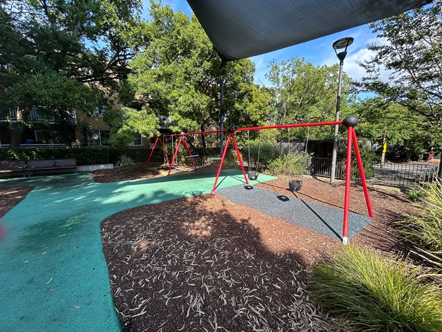 A playground with red swings on a colorful surface under trees, featuring a streetlamp and benches in the background.