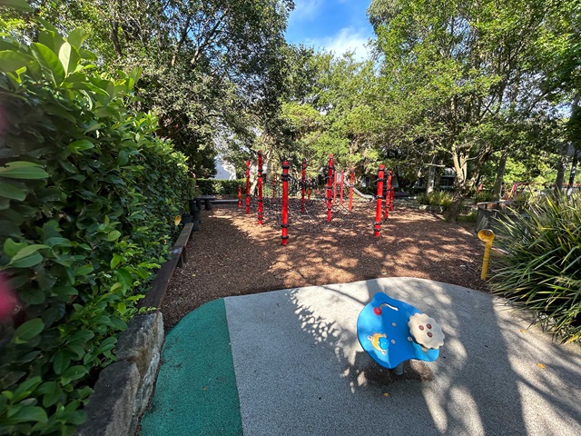 Children's playground with a blue spring rocker in the foreground and red climbing structures in a wood-chip area, surrounded by trees and greenery.