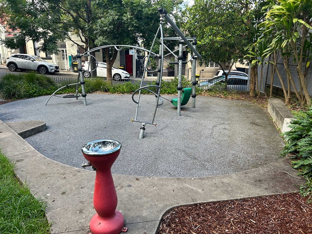 A small playground with climbing structures and a red water fountain. Trees and a parked car are in the background.