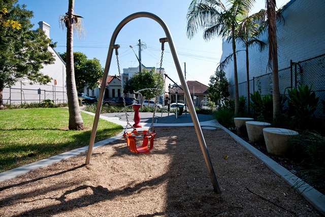 A playground swing set with two swings on a sandy area, surrounded by trees and buildings under a clear blue sky.