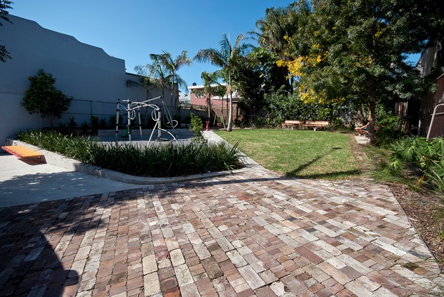 A brick-paved courtyard features outdoor gym equipment, a small lawn, trees, and a wooden bench under clear blue skies.