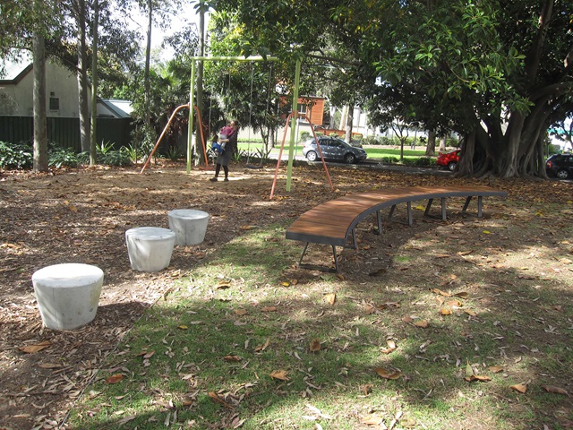 A person stands on a swing in a park surrounded by trees. Nearby are concrete stools and a curved bench on a grassy area. Cars are parked in the background.