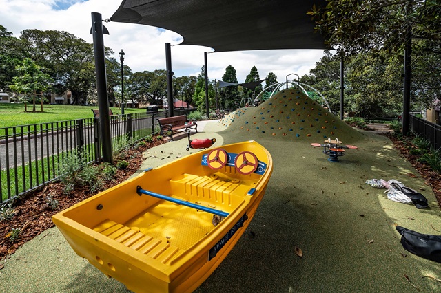 Playground with a yellow boat structure, climbing hill with footholds, and a small roundabout under a shaded canopy. Trees and a park bench are visible in the background.
