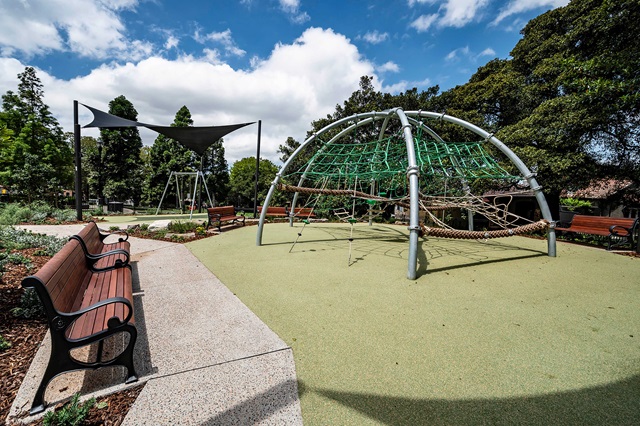 Playground with climbing structure, shaded swing set, benches, and trees under a partly cloudy sky.