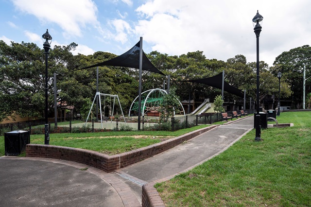 Playground with swings, a slide, and covered areas is surrounded by a fence. Pathways and streetlamps are visible, and trees are in the background.