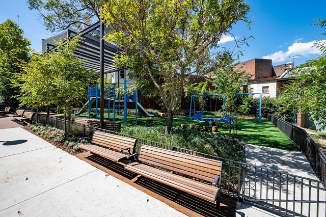 A small urban park with benches, green trees, a play area including slides, swings, and a shade structure on a sunny day.