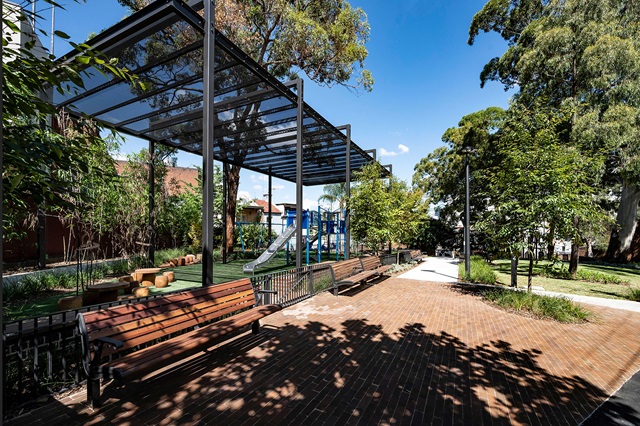 A park with benches, trees, and a covered area. A playground with slides is visible in the background.