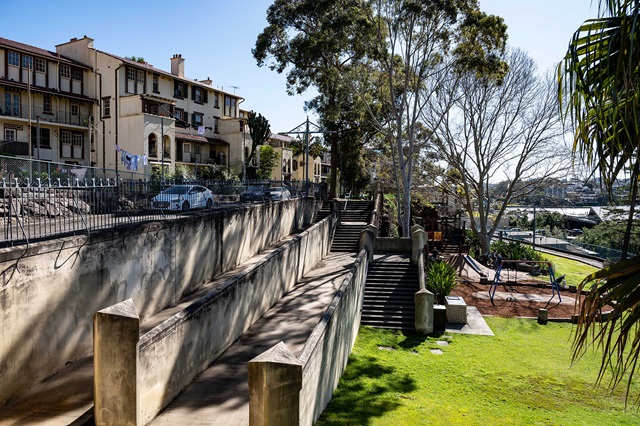 A row of residential buildings overlooks a staircase and playground, surrounded by trees and grass, under a clear sky.