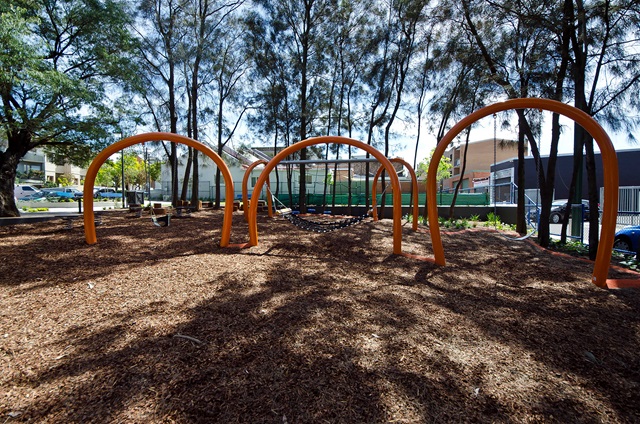 Playground with orange arches, a netted climbing structure, and wood chip ground cover, surrounded by trees and buildings.
