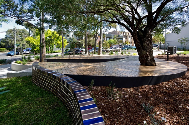A curved wooden platform surrounds a tree in a park setting, with a low brick wall in the foreground and parked cars visible in the background.