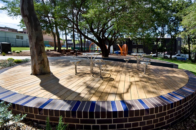 A circular wooden platform with tables and chairs is surrounded by a low brick wall and trees in an outdoor setting.