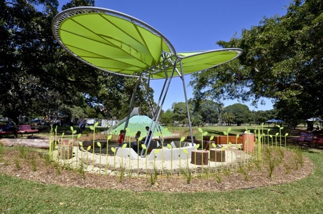 Playground with large green leaf-shaped shade structure, surrounded by grass, trees, and seating.