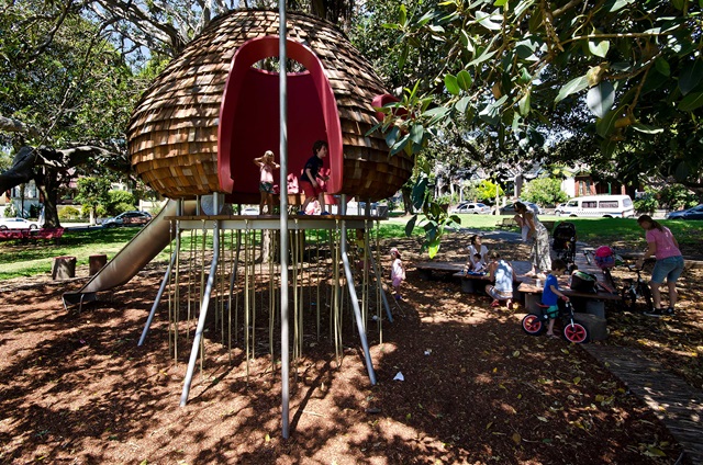 Children playing in a treehouse-themed playground structure with a slide, surrounded by trees and parents nearby.