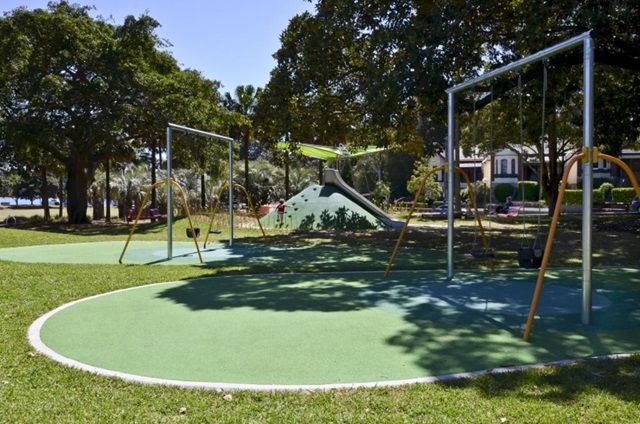 Playground scene with three empty swings on green turf, a climbing structure in the background, surrounded by trees and grass.