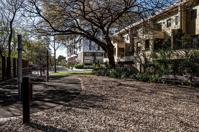 A playground with woodchip ground cover, a climbing structure, and benches, surrounded by trees and residential buildings on a sunny day.