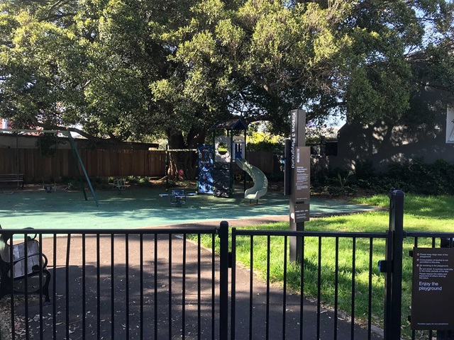 A playground with a slide, swings, and climbing structure, surrounded by trees and enclosed by a black fence.