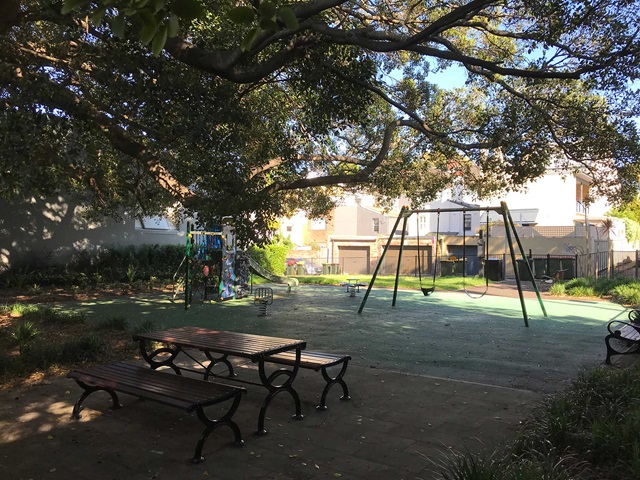 A small playground with a slide, swings, and benches under large trees. Nearby are a few houses under a bright blue sky.