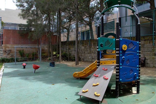 City playground with slide and green play area, surrounded by buildings and trees.